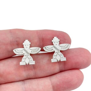 May include: A pair of silver-toned cufflinks. Each cufflink features a stylized figure with outstretched wings, a hat, and geometric patterns. The cufflinks are held in a hand against a white background.
