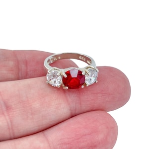 Könnte beinhalten: Ein silberner Ring mit drei Steinen. Der Mittelstein ist ein großer, oval geschliffener, leuchtend roter Edelstein, flankiert von zwei kleineren, runden, klaren Steinen. Der Ring wird in einer Hand vor weißem Hintergrund gehalten.