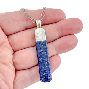 Peut inclure: Un pendentif rectangulaire composé d'une pierre de lapis-lazuli bleu profond avec des éclats dorés. La pierre est surmontée d'une bélière en métal argenté martelé et est suspendue à une fine chaîne à maillons ovales argentés.