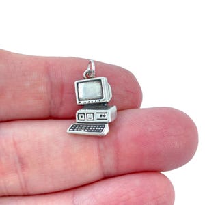 May include: A small, silver-colored computer charm with a monitor, keyboard, and base. The charm has a loop at the top for attaching to a necklace or bracelet. The computer charm is a miniature replica of a vintage desktop computer.