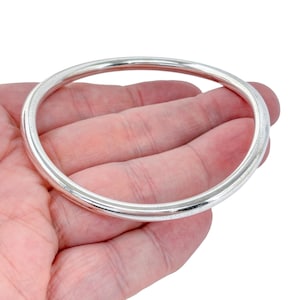 Puede incluir: Un brazalete plateado con un diseño liso y redondeado. El brazalete se sostiene en una mano sobre un fondo blanco. El brazalete es una pieza de joyería sencilla y elegante.