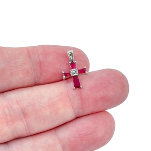 May include: A small, silver-toned cross pendant with a vertical bar and a horizontal bar. The cross features rectangular, deep red gemstones and a small, round, silver-toned accent at the center. The top of the cross has a small loop for a chain.
