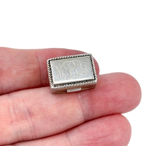 Puede incluir: Una pequeña caja rectangular de plata con tapa con bisagras. La tapa presenta un monograma grabado dentro de un marco rectangular con un borde en forma de cuerda. La caja se sostiene en una mano sobre un fondo blanco.