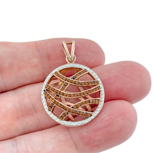 May include: A circular pendant featuring a rose gold-toned metal frame with an intricate, crisscrossing lattice design. The lattice is embellished with small, amber-colored stones, while the outer circular border is set with clear stones.