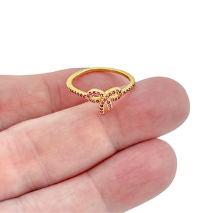 May include: A gold-toned ring featuring a central bow-shaped design adorned with small, vibrant pink stones. The band is thin and also encrusted with matching pink stones along the sides, creating a cohesive and detailed appearance for this jewelry piece.