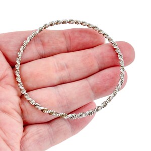 Puede incluir: Una pulsera rígida circular de metal plateado, que presenta una textura de cuerda trenzada continua en toda la banda. El diseño consiste en un solo hilo uniforme que forma un bucle completo con un acabado metálico pulido y brillante.