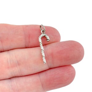 May include: A silver-toned metal charm shaped like a candy cane. The charm features a smooth, curved top and a straight, cylindrical body with a diagonal, spiraling groove pattern running along its length, ending in a small circular jump ring.
