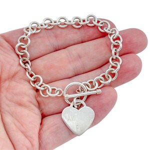 May include: A silver-toned chain link bracelet featuring a toggle clasp and a heart-shaped pendant. The pendant is engraved with the letters AM and PM stacked vertically. The bracelet is constructed from circular links connected in a continuous loop.