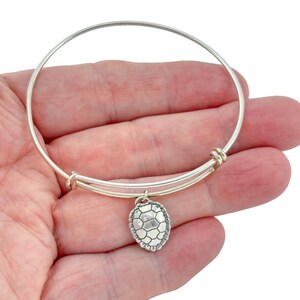 Puede incluir: Una pulsera tipo brazalete extensible de tono plateado con un dije colgante en forma de caparazón de tortuga. El dije tiene una superficie texturizada y segmentada, y la pulsera está construida con una banda de alambre circular.