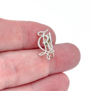 May include: A silver-colored metal charm featuring a stylized, cursive letter R with an intricate, looped decorative base. The piece has a polished finish and a small integrated loop at the top for attachment, showcasing a delicate, openwork design.