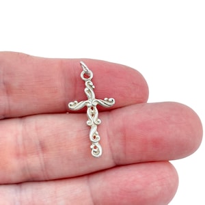 Peut inclure: Un petit pendentif en forme de croix argentée avec un motif orné de volutes. La croix a une boucle en haut pour une chaîne et est tenue dans la main d'une personne sur fond blanc. La croix mesure environ 3,8 cm de long.