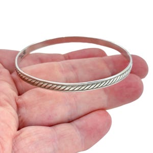 May include: A silver-toned bangle bracelet featuring a continuous twisted rope-like texture along the outer surface. The metal band is circular and polished, showcasing a repeating diagonal pattern that encircles the entire circumference of the piece.