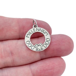 Peut inclure: Pendentif argenté avec le mot "MILLENNIUM" gravé sur le bord extérieur. Le pendentif présente un motif en forme de cœur au centre et est conçu pour être porté comme un charme de collier.