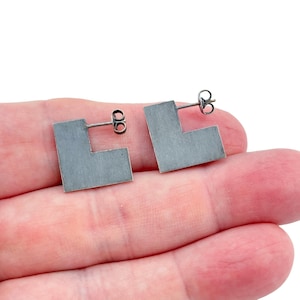 May include: A pair of silver-toned, L-shaped stud earrings. The earrings have a brushed metal finish and a simple, geometric design. The posts are visible, and the earrings are displayed on a light-colored background.