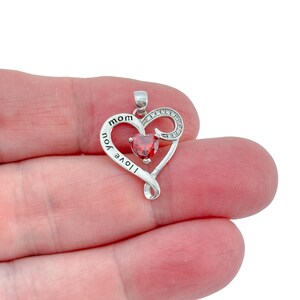 May include: A silver-toned heart-shaped pendant featuring the engraved text I love you mom. The design includes a central red heart-shaped gemstone and a curved side accent embellished with small clear stones, finished with a metal bail at the top.