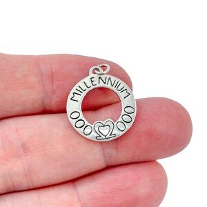 Op de afbeelding: Een zilverkleurige bedel met het woord "MILLENNIUM" in een cirkel. De bedel heeft een hartvorm en drie cirkels aan elke kant. Het wordt in de hand van een persoon gehouden.