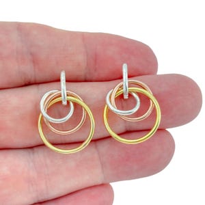 Pode incluir: Um par de brincos pendentes com três anéis circulares entrelaçados em tons de prata, ouro rosa e ouro amarelo. Cada brinco consiste num pequeno aro superior prateado ligado a um anel de ouro rosa e a um anel de ouro amarelo maiores.