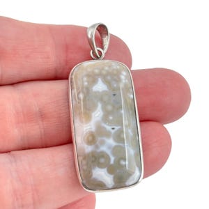 May include: A rectangular pendant featuring a polished, smooth stone with a natural pattern of tan and white circular orbs. The stone is encased in a simple silver-toned metal frame with a matching silver-toned bail at the top for hanging.