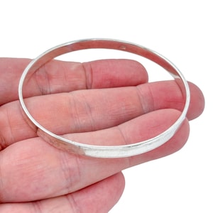 May include: A smooth, polished silver-toned bangle bracelet with a simple, circular design. The metal band features a reflective surface and a consistent, uniform width throughout its entire circumference, showcasing a clean and unadorned aesthetic.