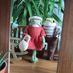 May include: Two crocheted frog figurines standing in a doorway. The frog on the left is wearing a red dress and carrying a white crocheted bag. The frog on the right is wearing a brown and white striped sweater and brown pants.