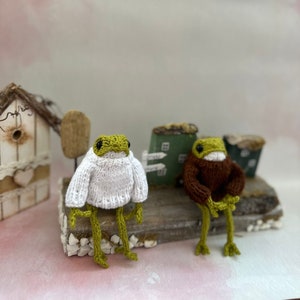 May include: Two knitted frog figurines wearing sweaters, one white and one brown, sitting on a wooden surface with a small house in the background.