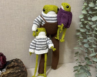 Op de afbeelding: Drie gebreide kikker knuffels die verschillende outfits dragen. De grootste kikker draagt een bruine overall en een wit-grijze gestreepte trui. De middelste kikker draagt een paarse trui. De kleinste kikker draagt een wit-grijze gestreepte trui en een bruine broek.