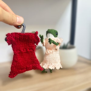 May include: A green frog wearing a pink knitted dress with a ruffled hem, holding a red knitted dress on a hanger.