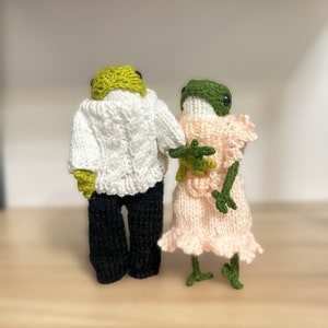 May include: Two crocheted frog plushies, one wearing a white sweater and black pants, the other wearing a pink dress. The frogs are standing on a wooden surface.