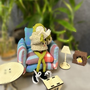 May include: A small knitted figure wearing a sweater and pants sits in a plaid armchair reading a book. The figure is sitting in a miniature living room setting with a small table, lamp, and fireplace.