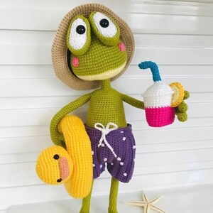 Puede incluir: Una rana de crochet verde que lleva un sombrero de paja, pantalones de baño de lunares blancos y morados y sostiene un flotador inflable amarillo con un pato amarillo. La rana también sostiene una taza de crochet rosa y blanca con una pajita azul.