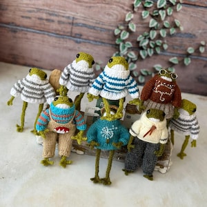 May include: A collection of handmade frog figurines, each wearing a unique knitted outfit. The frogs have green heads and long legs, with outfits in various colors and patterns, including sweaters, pants, and scarves. The background is a wooden surface.