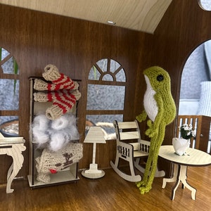 May include: A green knitted frog figurine standing next to a rocking chair and a small wooden table with a white flower arrangement in a white pot. The frog is wearing a white and red striped sweater. The scene is set in a miniature wooden dollhouse.
