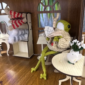 May include: A green knitted frog wearing a beige knitted sweater sits in a chair reading a book. The book has the text "Frog's cup cake" written on it. The frog is in a miniature dollhouse setting with a small table and a vase of white flowers.