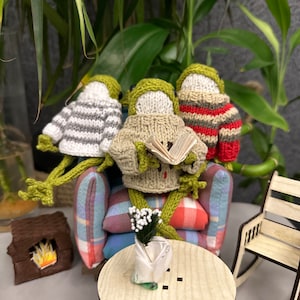 May include: Three knitted frog figurines sitting on a plaid couch with a book, a small table with a vase of white flowers, and a rocking chair in the background.