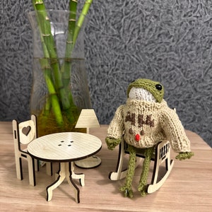 May include: A green knitted frog wearing a beige sweater with a red pom-pom sits in a wooden rocking chair. A small wooden table and chair are in the background.