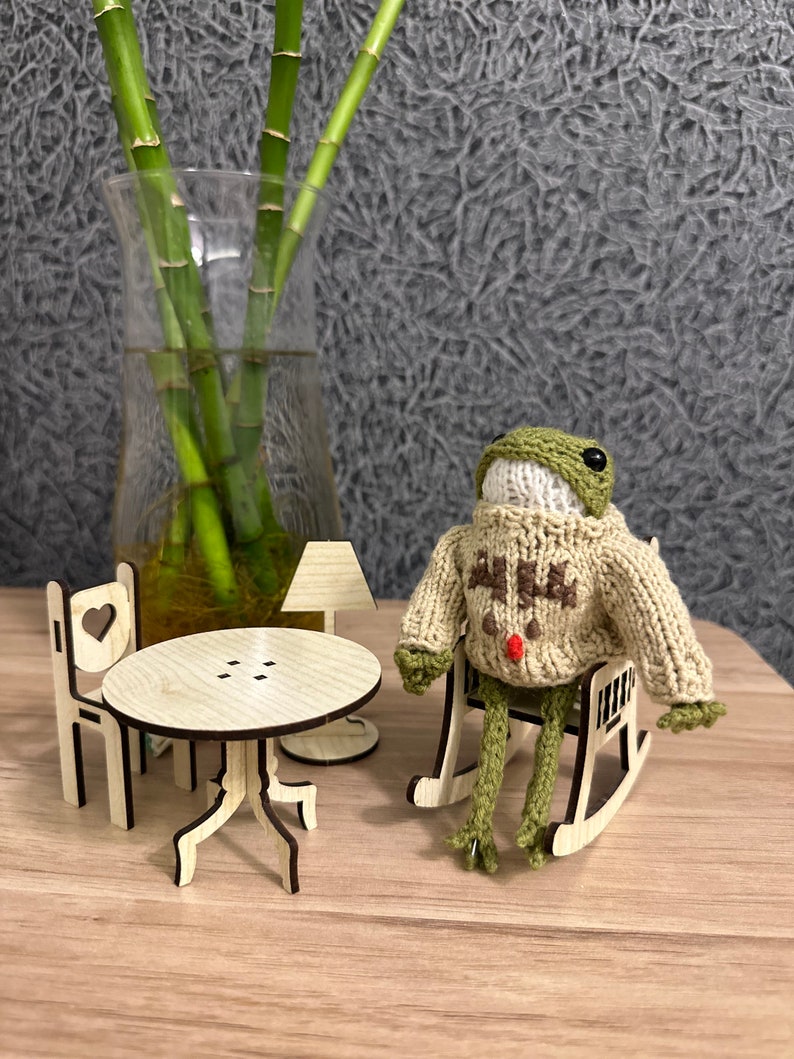 May include: A green knitted frog wearing a beige sweater with a red pom-pom sits in a wooden rocking chair. A small wooden table and chair are in the background.