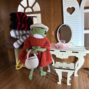 Könnte beinhalten: Ein grüner Frosch, der ein rotes Strickkleid und eine weiße Strickmütze trägt, hält eine weiße Stricktasche. Der Frosch steht in einem Puppenhaus-Setting mit einem Schminktisch und einem Hocker.