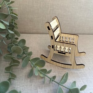 May include: A miniature wooden rocking chair with a light brown finish. The chair has a slatted back and a simple design.