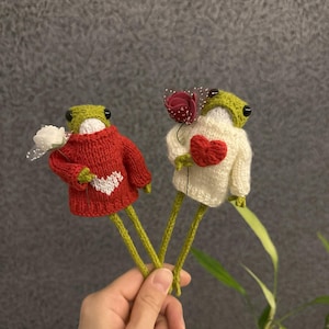 May include: Two knitted frog figurines wearing sweaters and holding flowers. The frog on the left is wearing a red sweater with a white heart and holding a white flower. The frog on the right is wearing a white sweater with a red heart and holding a red flower.