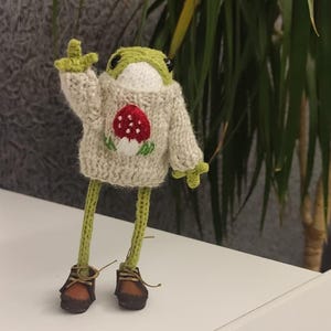Op de afbeelding: Een handgemaakte gehaakte kikkerpop met een groene kop en ledematen, gekleed in een beige trui met een rood paddestoelontwerp. De kikker heeft bruine schoenen en heft een arm op.