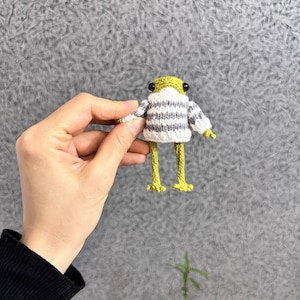 May include: A small, handmade frog figurine with a green body and yellow legs. The frog is wearing a white and gray striped sweater. The frog has black eyes and is held in a person's hand.