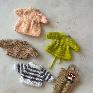 May include: A collection of miniature knitted doll clothes in various colors. Includes a peach-colored dress, a brown sweater, a green top, a striped sweater, and tan pants with a mushroom design. The items are arranged on a light-colored surface.
