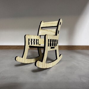 May include: A miniature wooden rocking chair with a black outline. The chair has a slatted back and seat.