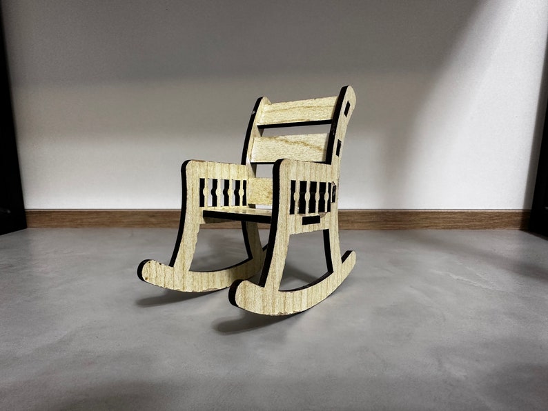 May include: A miniature wooden rocking chair with a black outline. The chair has a slatted back and seat.