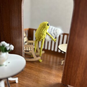 May include: A green knitted frog doll sitting in a rocking chair on a wooden porch. The frog is looking out of the doorway.
