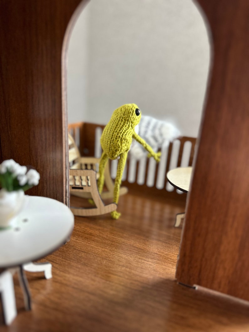 May include: A green knitted frog doll sitting in a rocking chair on a wooden porch. The frog is looking out of the doorway.