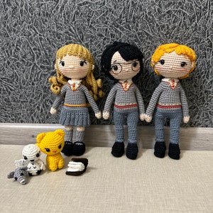 May include: Three crocheted dolls dressed in Gryffindor robes, standing in front of a gray textured wall. The dolls are a girl with blonde hair, a boy with black hair and glasses, and a boy with blonde hair. There are also two small crocheted animals in front of the dolls.