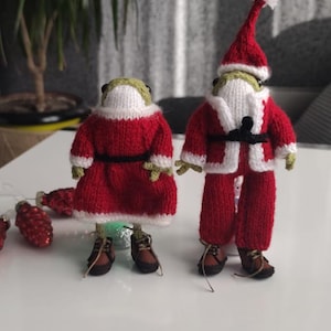 Puede incluir: Dos figuras de rana tejidas vestidas con trajes de Papá Noel. Las ranas visten trajes rojos con ribetes blancos, cinturones negros y zapatos marrones. Una tiene un gorro de Papá Noel. Hay adornos navideños esparcidos cerca.