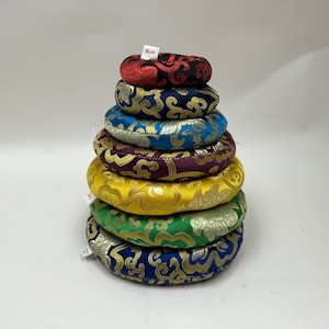 以下が含まれることがあります： 赤、青、緑、黄、紫など、さまざまな色と模様の丸い瞑想クッションが7つ積み重ねられています。クッションは複雑なデザインの布地で作られており、ピラミッド型に積み重ねられています。一番上のクッションには、「10 cm」と書かれた小さな白いタグが付いています。