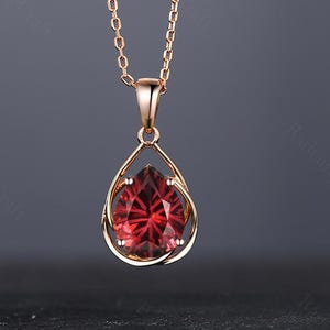 May include: A rose gold necklace with a teardrop pendant. The pendant holds a faceted, deep red gemstone in an openwork frame. The fine chain enhances the pendant's elegant design. The necklace is a piece of jewellery.